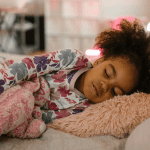 A small child sleeping on a fluffy pink cushion with a pink and white blanket.