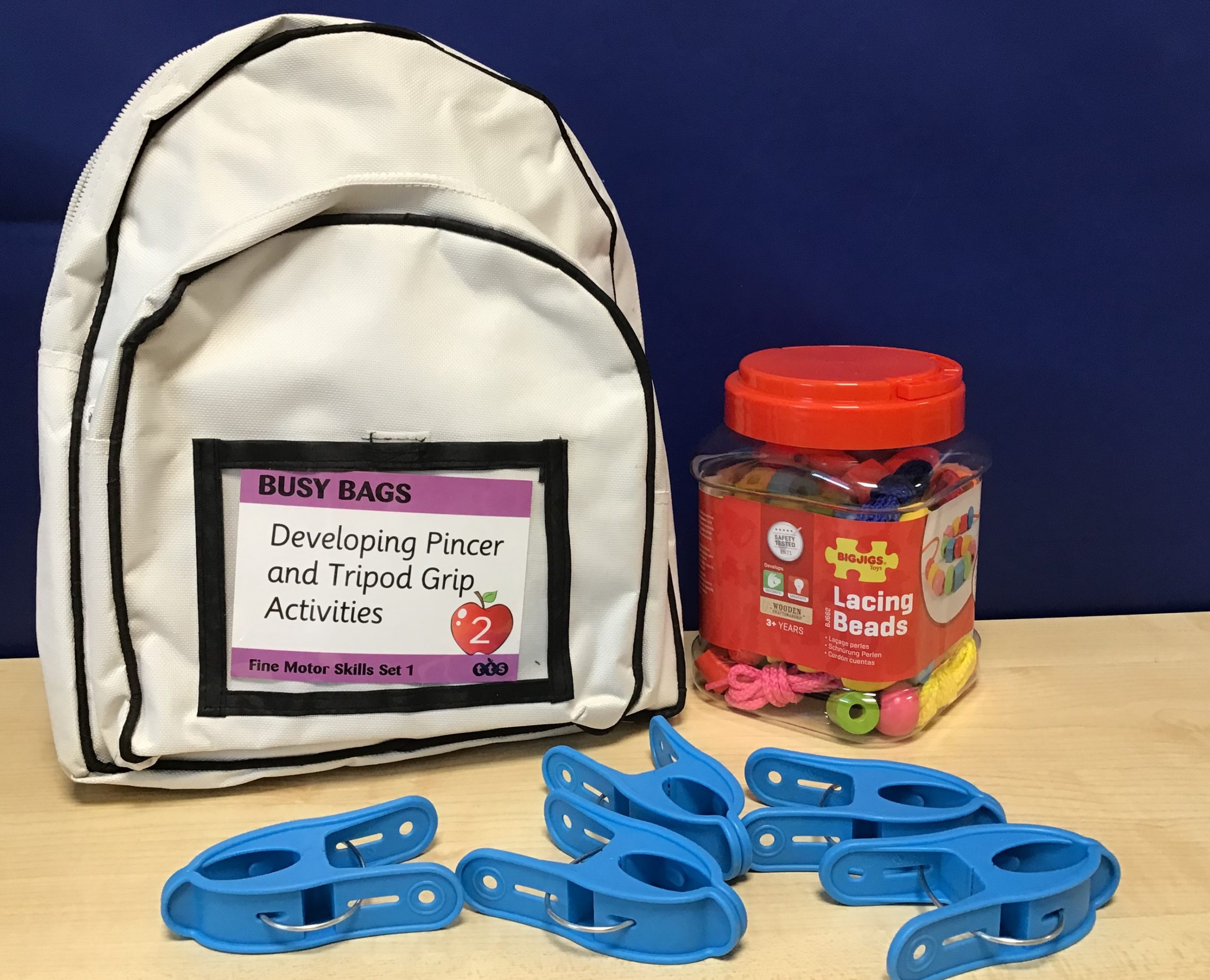 Busy Bags – Fine Motor Skills. White backpack with a box of colourful lacing beads and blue pincer clips.