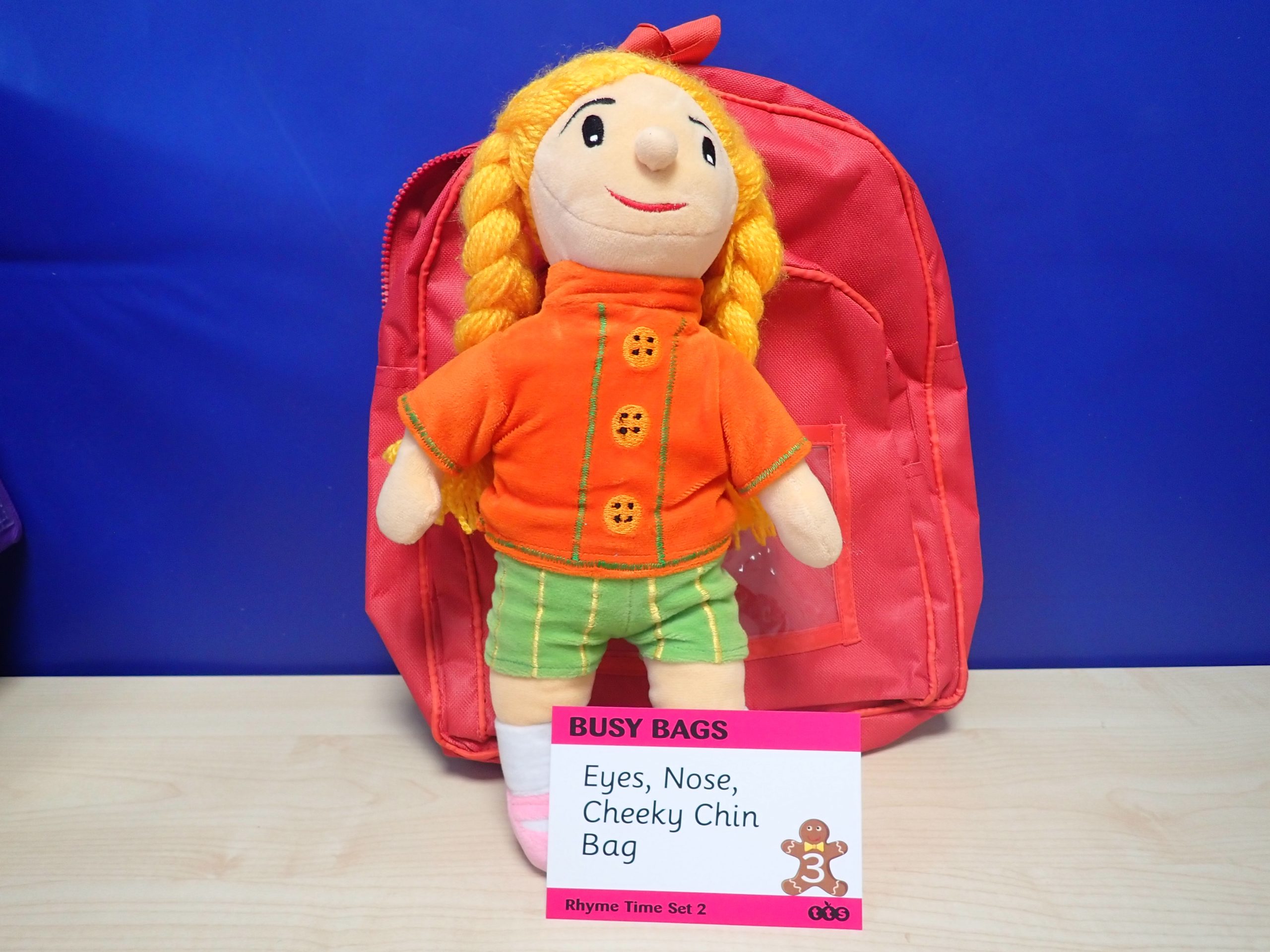 Rhyme time bag – Eyes, Nose, Cheeky Chin. Red back pack with a soft toy of a young girl with braided yellow hair. The girl is wearing an orange top, green shorts and pink shoes with white socks.
